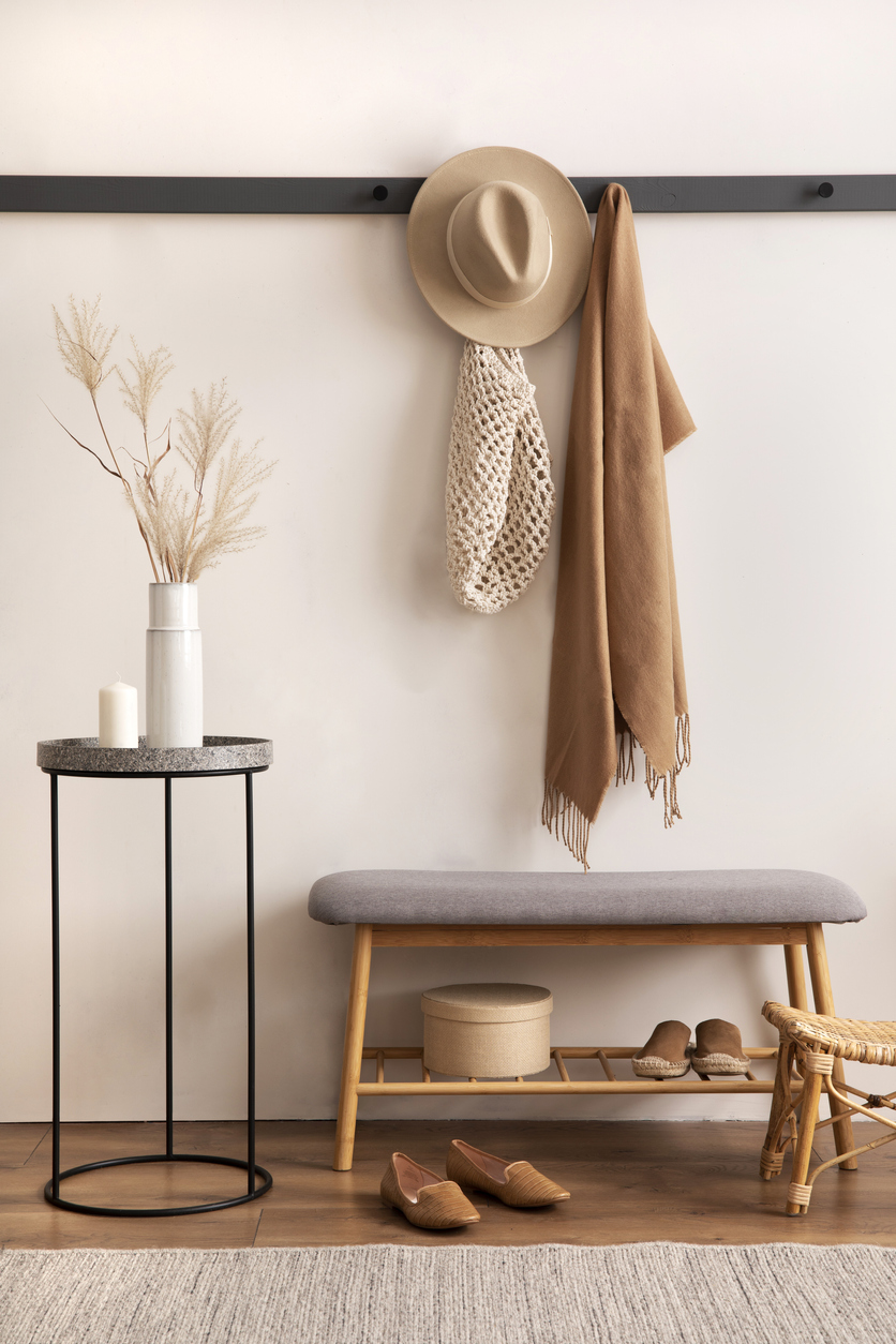 Warm and cozy interior design of bright entryway with gray bench, black hanger, brown scarf, beige hat, round coffee table, white vase with dried flowers, shoes, woven shopping bag and personal accessories. Home decor. Template.