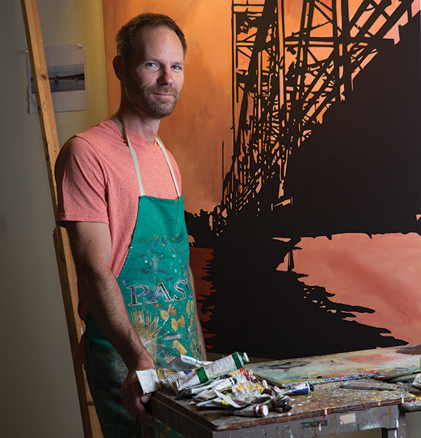 Artist Michael Slagle stands behind a table with painting supplies.