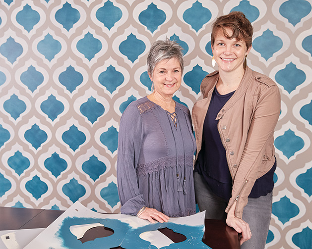 The two women behind Gypsy Mint Stencil Co. pose in front of some stencil work.