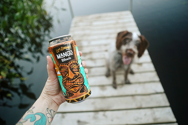A person holds a can of Lift Bridge Mango Blonde while a dog sitting on a dock watches.