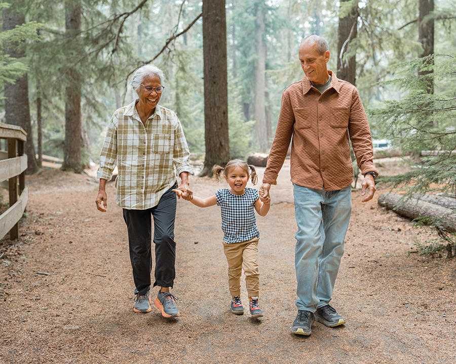 An active and healthy Pacific Islander senior woman and her handsome Caucasian husband hold hands with their cute Eurasian three year old granddaughter and smile while hiking together through a forest in the Pacific Northwest region of the United States.