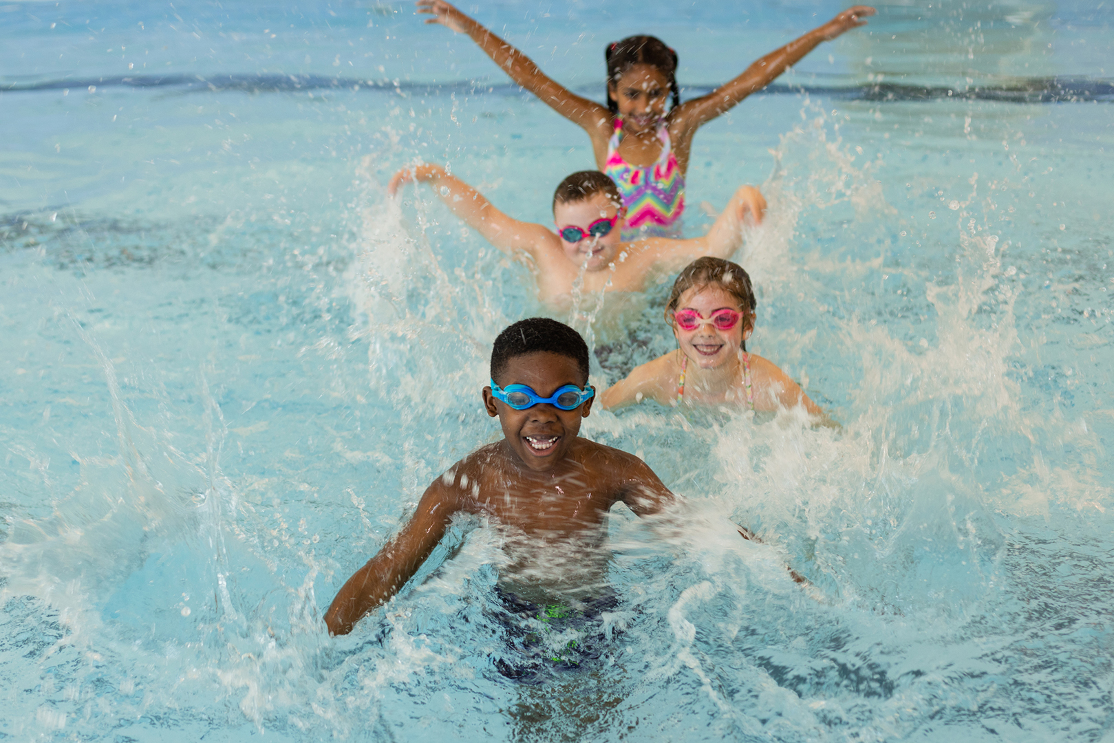 A group of children playing in the swimming pool in Boldon, North East England. They are standing behind each other and making a splash in the water with their arms while looking at the camera and wearing goggles.